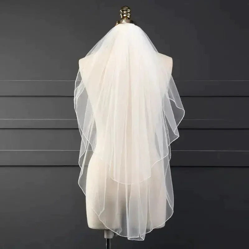 Ethereal double-tiered wedding veil by MANDOTOS with delicate lace edges and flowing tulle, displayed on a neutral background.