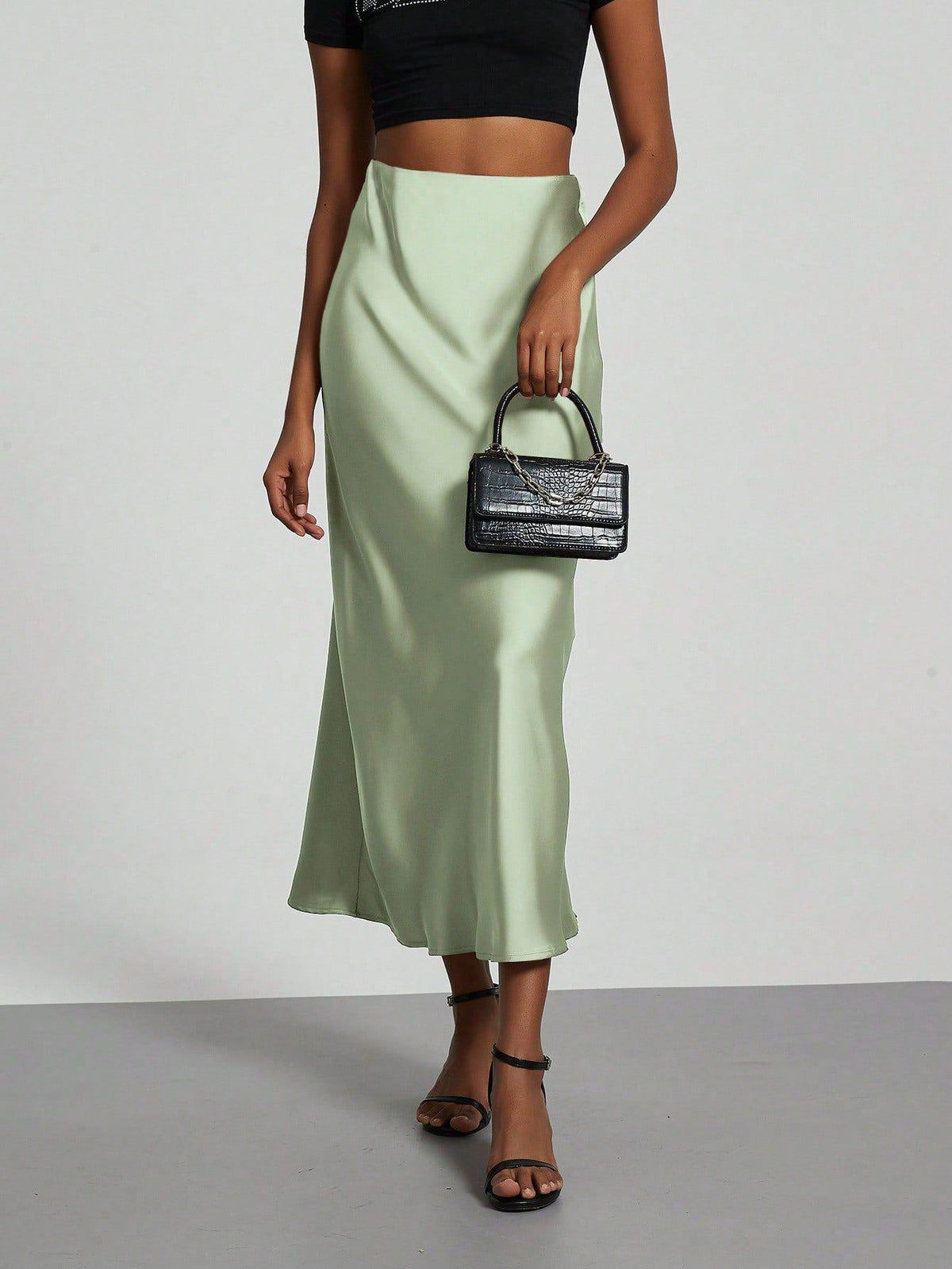 Woman wearing an elegant light green satin midi skirt, black crop top, and black handbag.